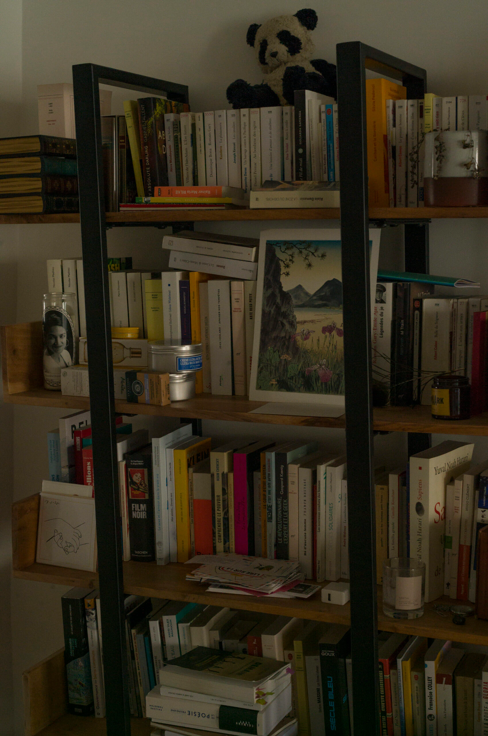Étagère en bois foncé chargée de livres — Kundera, Camus, Silvia Federici, Yuval Noah Harari — avec une peluche panda et une petite illustration japonisante
